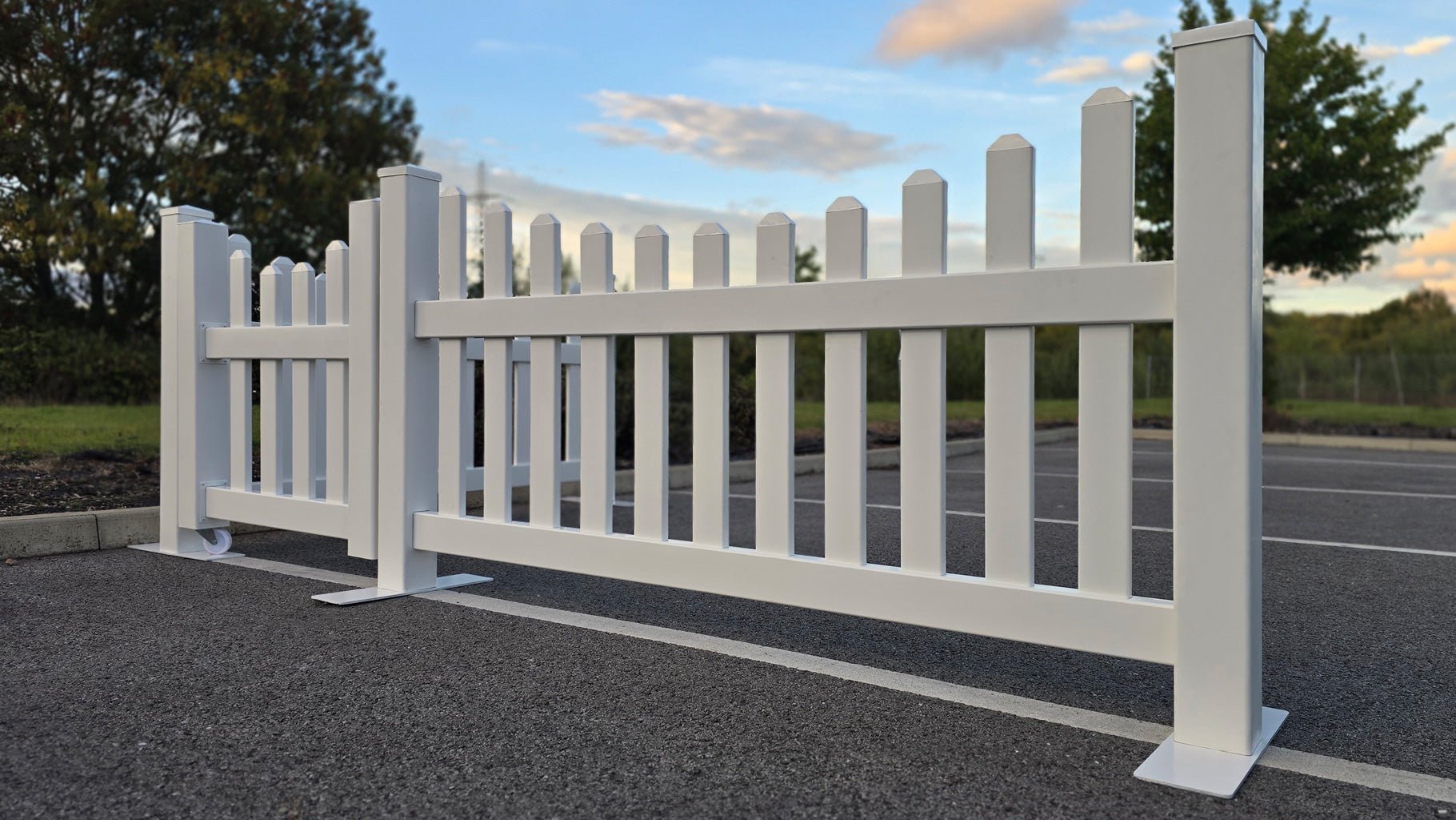 A portable scalloped vinyl picket fence set with a 3ft gate, shown on a car park surface with steel base plates for stability.