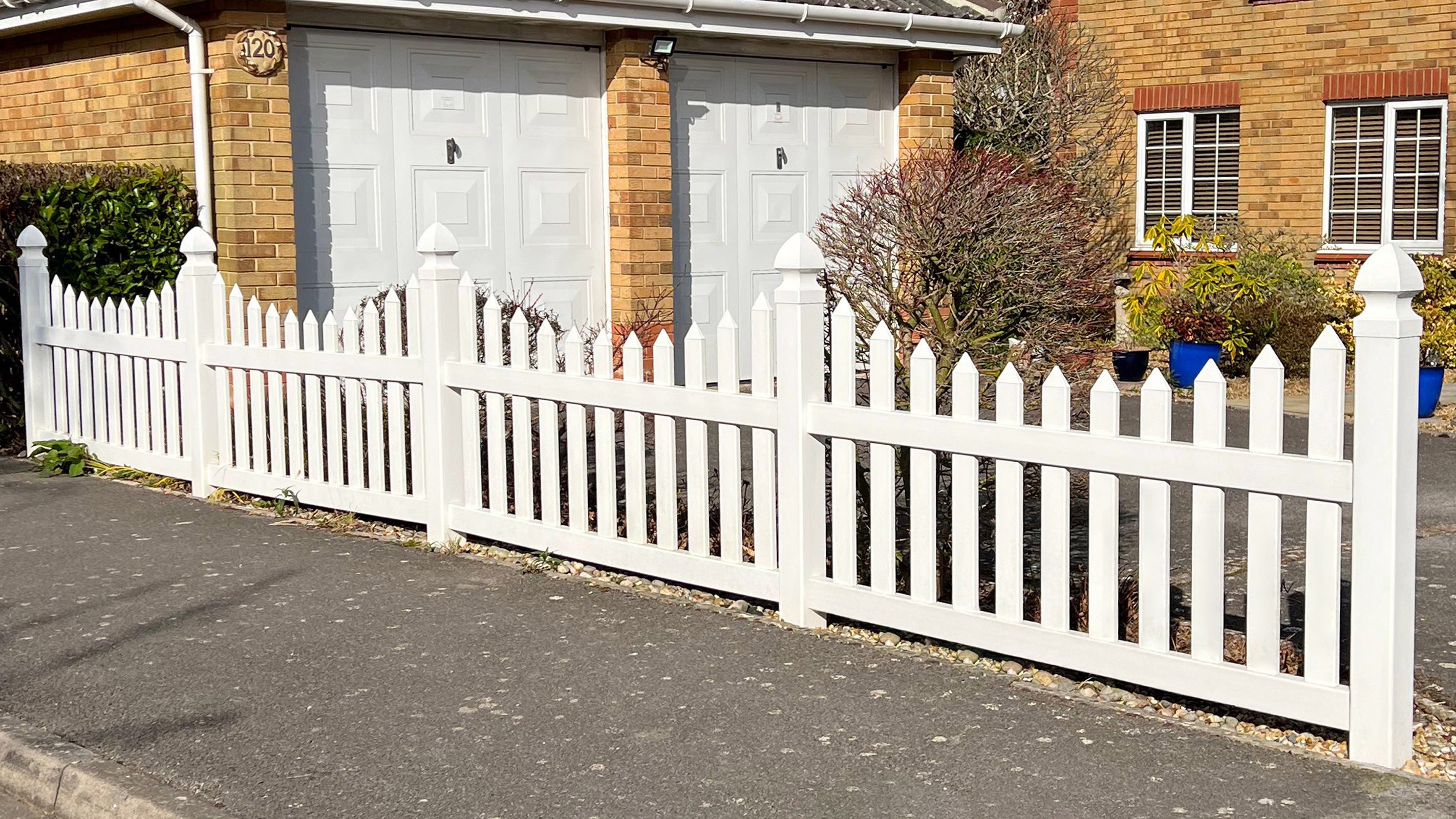 White scalloped picket fence in front of a house with a driveway.