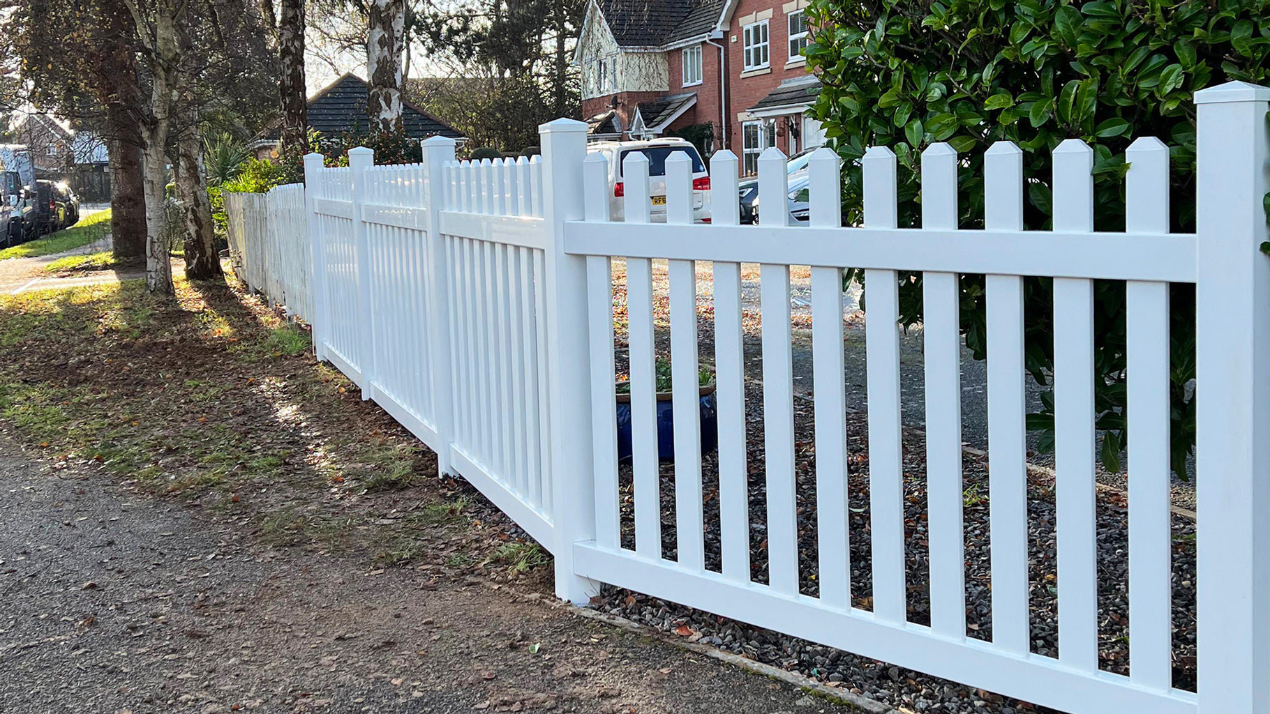 White picket fence along a residential street with houses and cars in the background by Simple Fencing.