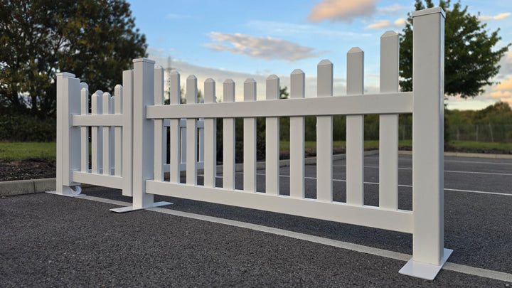 A portable scalloped vinyl picket fence set with a 3ft gate, shown on a car park surface with steel base plates for stability.